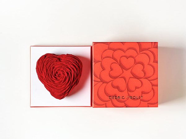 A heart-shaped red pastry, intricately designed with rose-like swirls, is elegantly presented in a luxurious red box adorned with a heart motif and the name 