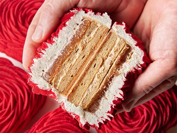 A close-up of the heart-shaped pastry sliced in half, revealing its delicate layers of sponge, cream, and caramel-like filling, encased in a vibrant red, textured exterior.