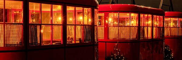 A row of red cable car cabins glowing warmly with soft interior lighting, adorned with checkered curtains and festive decor, set against a nighttime cityscape.