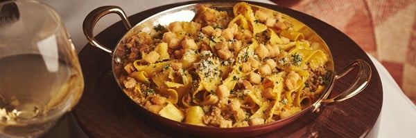 A luxurious pasta dish with golden ribbons of pappardelle, generously coated in a rich, savoury sauce, topped with crispy croutons and freshly grated cheese, served in an elegant brass-handled pan.