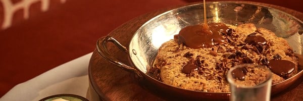 A decadent, freshly baked cookie dessert, studded with nuts and chocolate, drizzled with silky caramel sauce, served in a gleaming copper pan for a rustic yet indulgent presentation.