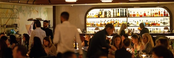 A vibrant, upscale restaurant bar with patrons enjoying a lively evening atmosphere, featuring a well-stocked illuminated back bar and waitstaff in motion, framed by soft, elegant decor.