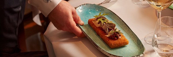 A meticulously plated dish featuring a golden, crisp base topped with delicate truffle shavings, fresh arugula, and paper-thin cheese, presented on a striking turquoise plate.