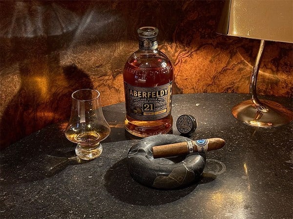 A refined scene featuring Aberfeldy 21 whisky, a cigar in a marble ashtray, a glass, and a golden lamp.