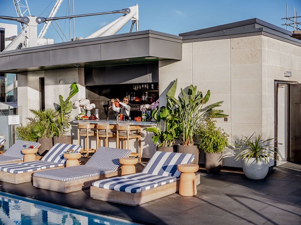 View of the Rooftop Bar, with blue & white striped sunloungers in a row/ Behind, is a man behind the bar, shaking a cocktail.