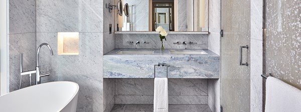 Marble bathroom with bath on the left, a shower on the right and a double basin in the centre of the room with a vase of white roses.