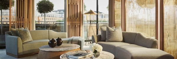 An inviting living space with curved neutral-toned sofas, marble-topped coffee tables, and floor-to-ceiling glass doors framed by bespoke wooden screens, opening onto a private terrace.