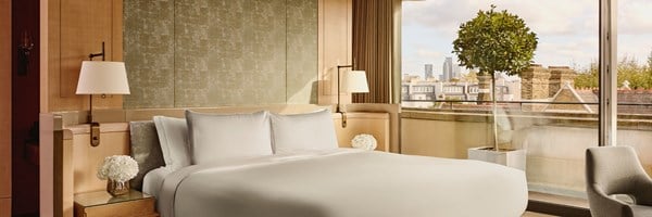 A tranquil bedroom sanctuary with a plush king-size bed, textured headboard, warm ambient lighting, and a glass wall revealing rooftop views of London’s skyline.