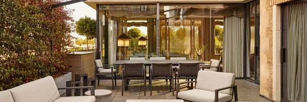 An expansive outdoor dining terrace with sleek modern furniture, soft-toned seating, and sculptural topiary, seamlessly connected to the interior via full-height glass doors.