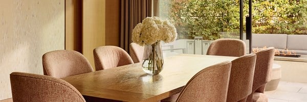 An elegant indoor dining space with a natural wood table, upholstered blush-toned chairs, and a crystal-clear vase of fresh hydrangeas, looking out to a landscaped terrace with fire feature.