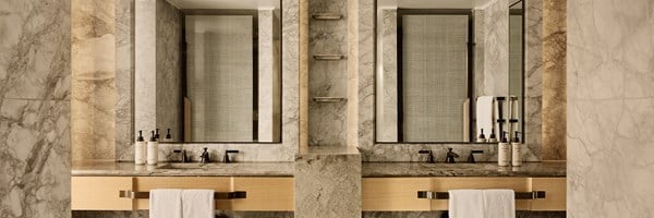 A sophisticated bathroom with twin marble vanities, brushed metal fixtures, and full-height mirrors, all framed in rich natural stone and soft lighting.