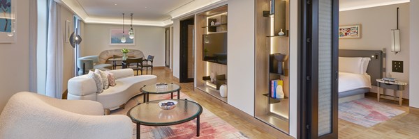 A modern luxury hotel suite featuring a stylish living area with curved seating, a dining table, built-in shelves, and a glimpse of the adjoining bedroom.