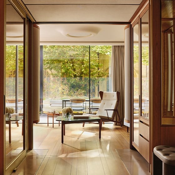 Warm, wood-accented living space with a modern lounge chair and glass coffee table, opening onto a sunlit terrace framed by trees in early autumn colours.