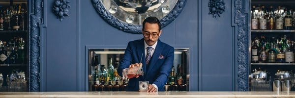 A dapper bartender in a tailored blue suit preparing a classic cocktail in an elegant bar with intricate decor.